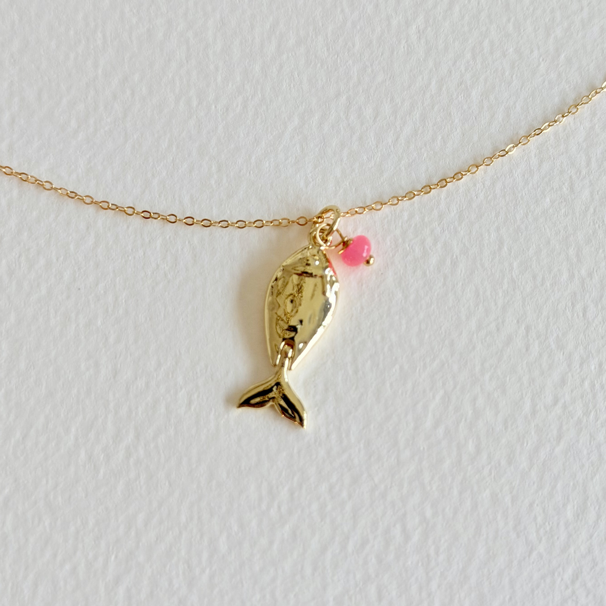 Gold fish-shaped pendant with a pink heart on a gold chain against a light gray background