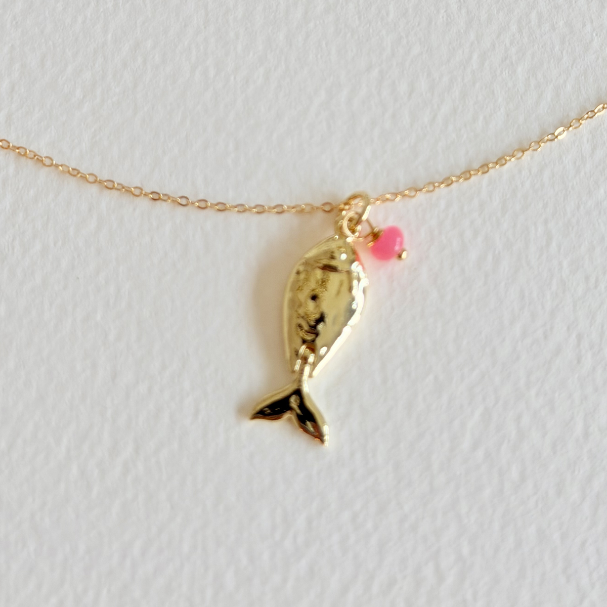 Gold fish-shaped pendant on a chain with a pink bead against a light gray background