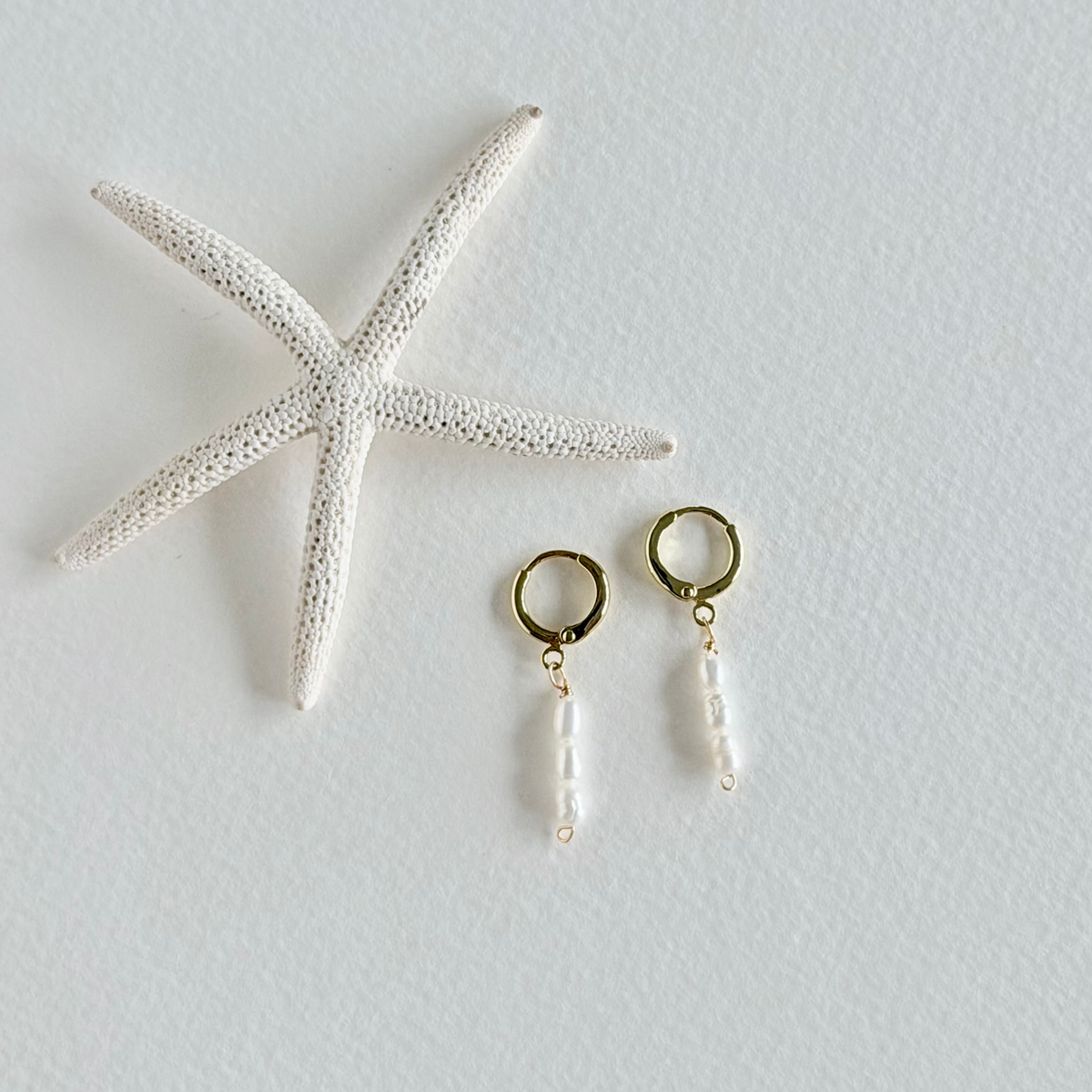 Pair of gold hoop earrings with pearl drops next to a starfish on a light gray background