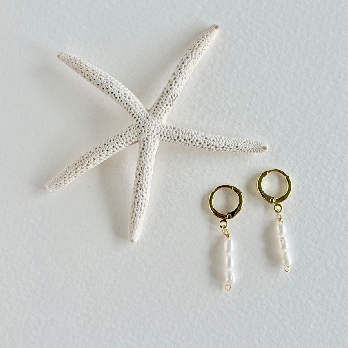 Gold hoop earrings with pearl drops next to a starfish on a light gray background