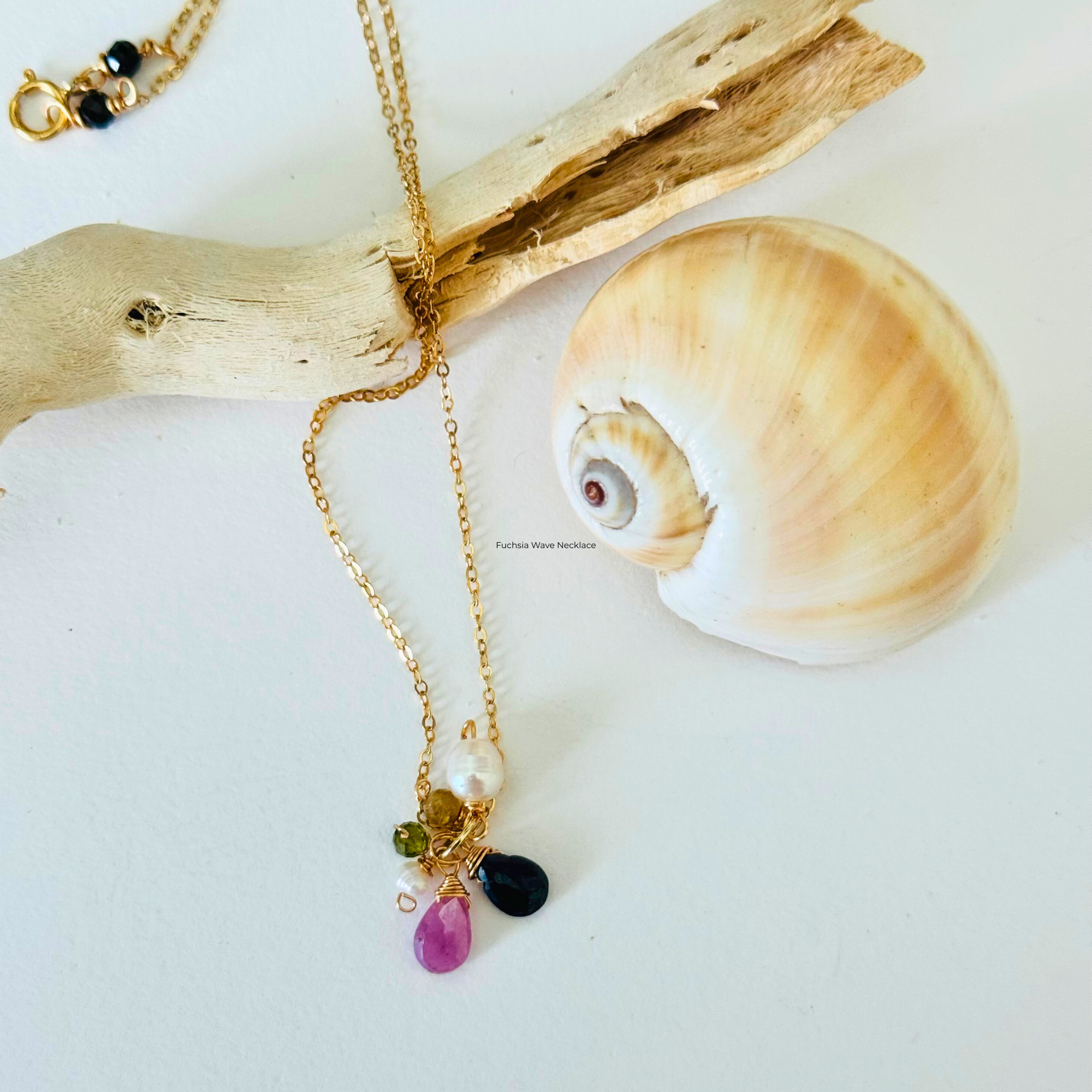 Pink sapphire, dark blue apatite, peridot, and freshwater pearl stones at end of handcrafted 14k gold filled wire necklace chain. Displayed draped over a bleached driftwood branch on white background framed by a creamy spiral seashell.