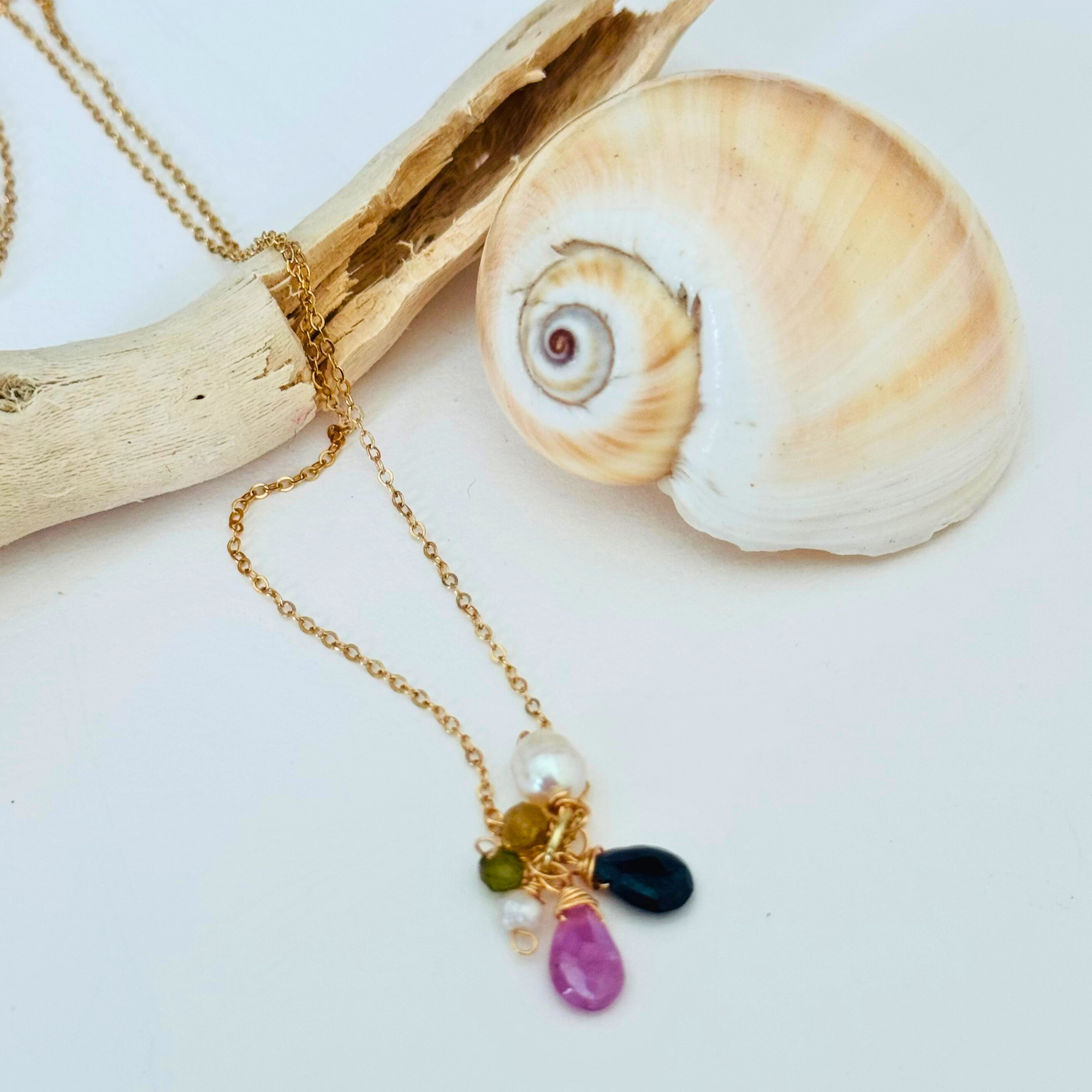 Pink sapphire, dark blue apatite, peridot, and freshwater pearl stones at end of handcrafted 14k gold filled wire necklace chain. Displayed draped over a bleached driftwood branch on white background framed by a creamy spiral seashell.