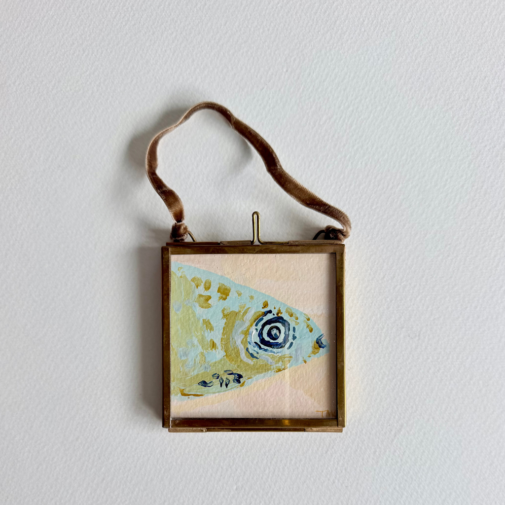 A square framed original painting featuring a stylized fish illustration in light blue, gold, and dark blue, suspended by a brown leather strap against a white wall.
