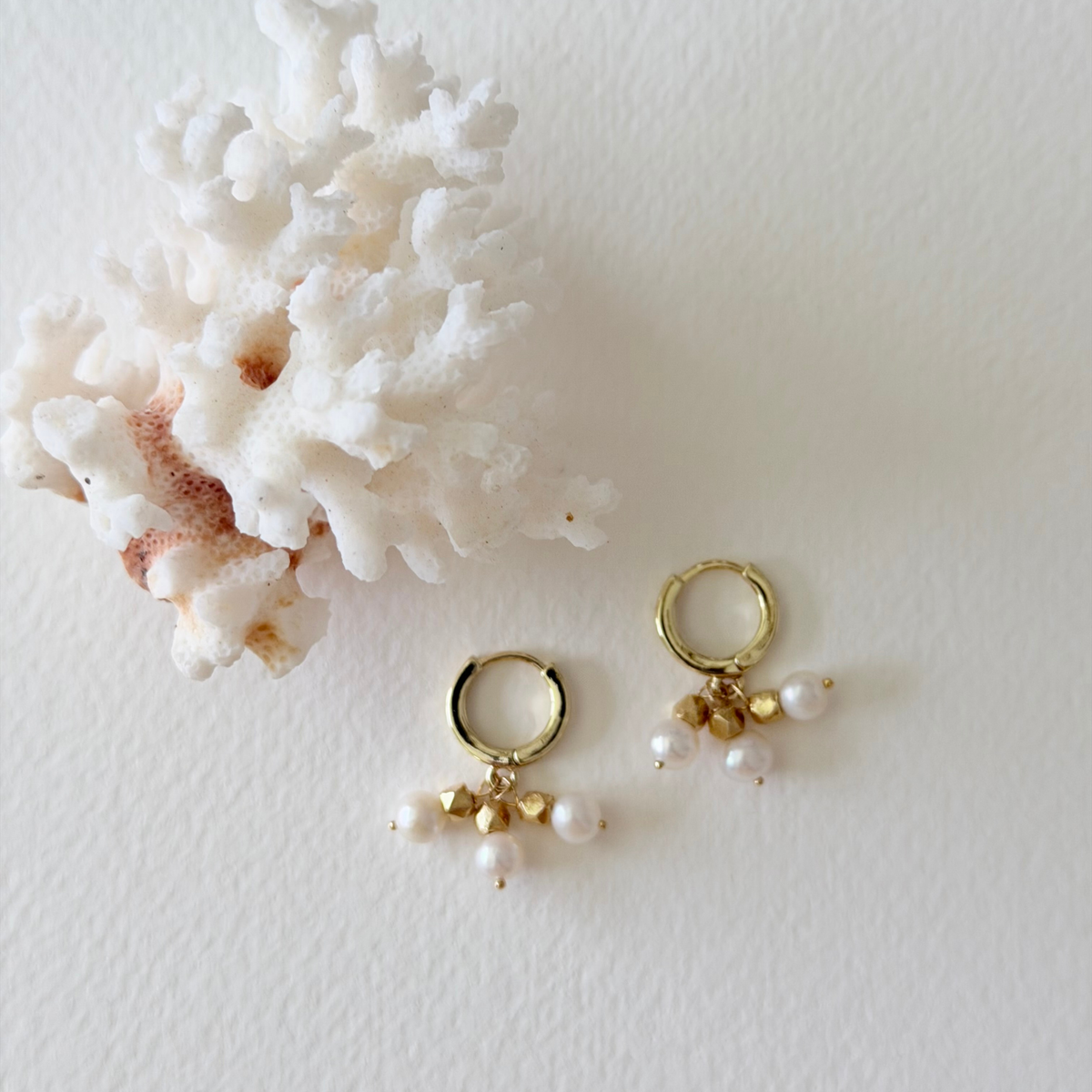 Gold hoop earrings with pearl charms on a white surface with coral.