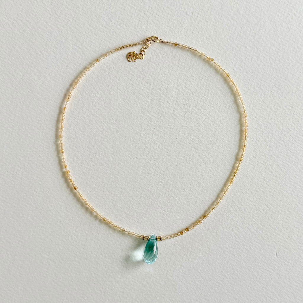 A delicate gold citrine beaded necklace with a small blue aquamarine teardrop pendant, displayed on a textured off-white background.
