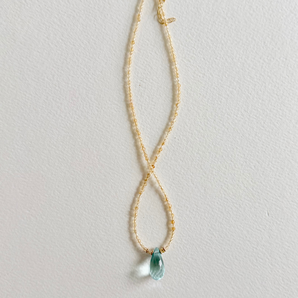 A delicate gold citrine beaded necklace with a small, faceted light blue teardrop aquamarine pendant, displayed against a textured off-white background.