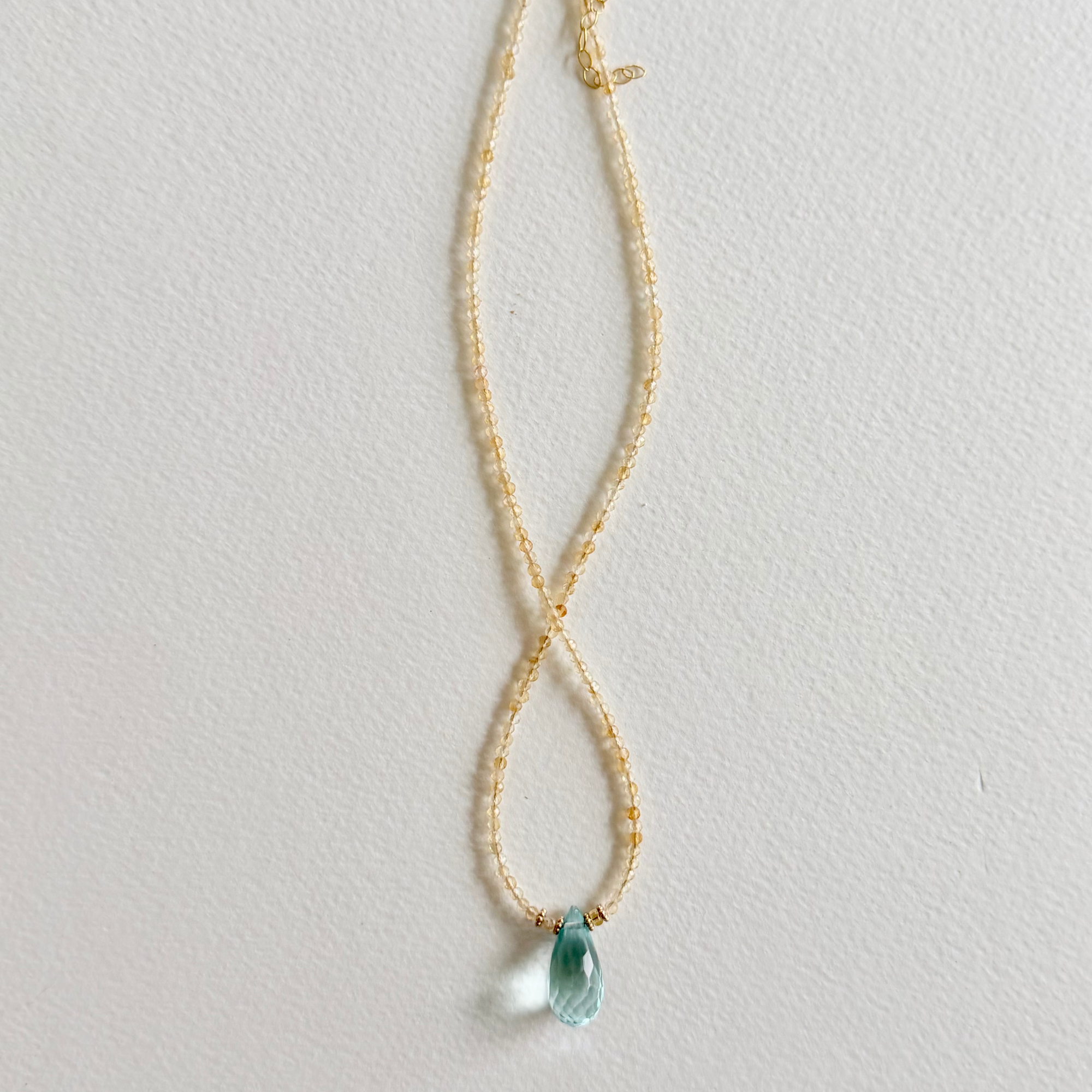A delicate gold citrine beaded necklace with a small, faceted light blue teardrop aquamarine pendant, displayed against a textured off-white background.