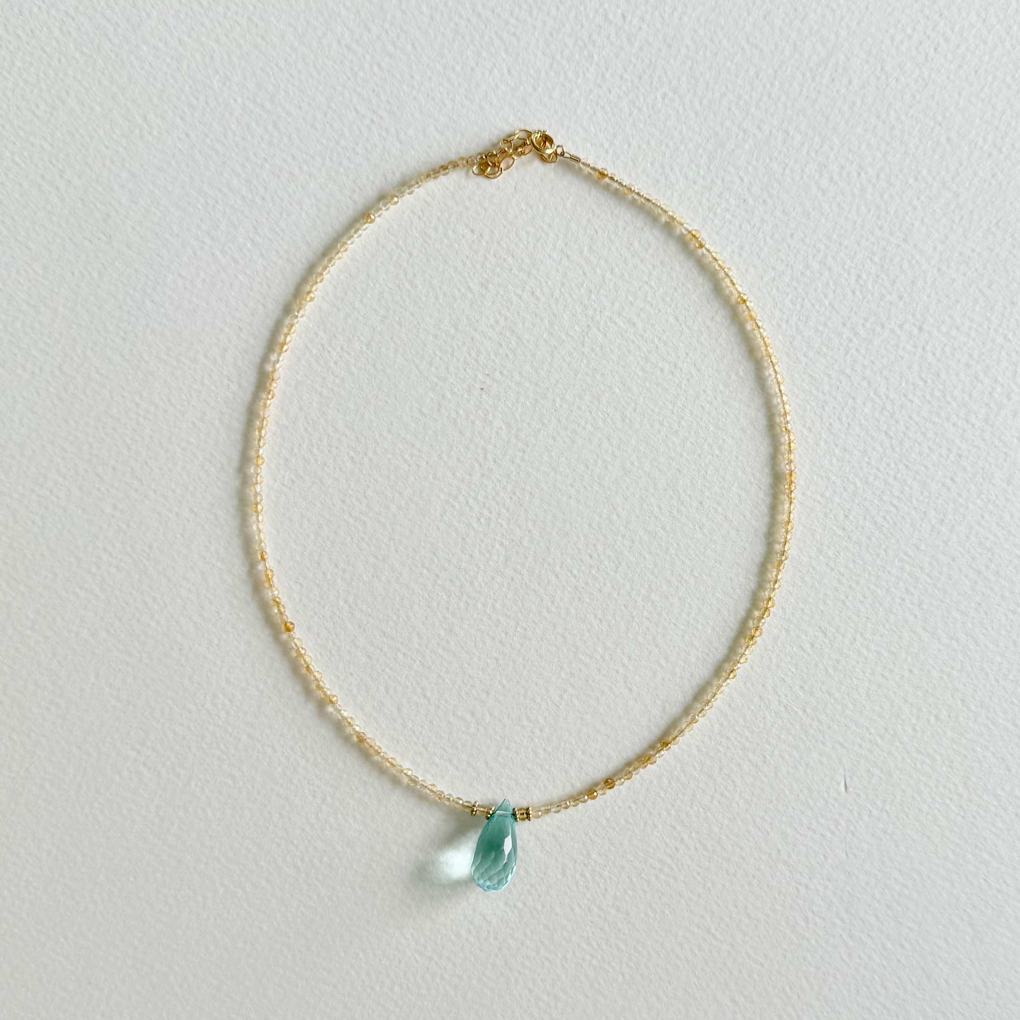 A delicate gold citrine beaded necklace with a small, teardrop-shaped light blue aquamarine pendant, displayed on a textured off-white background.