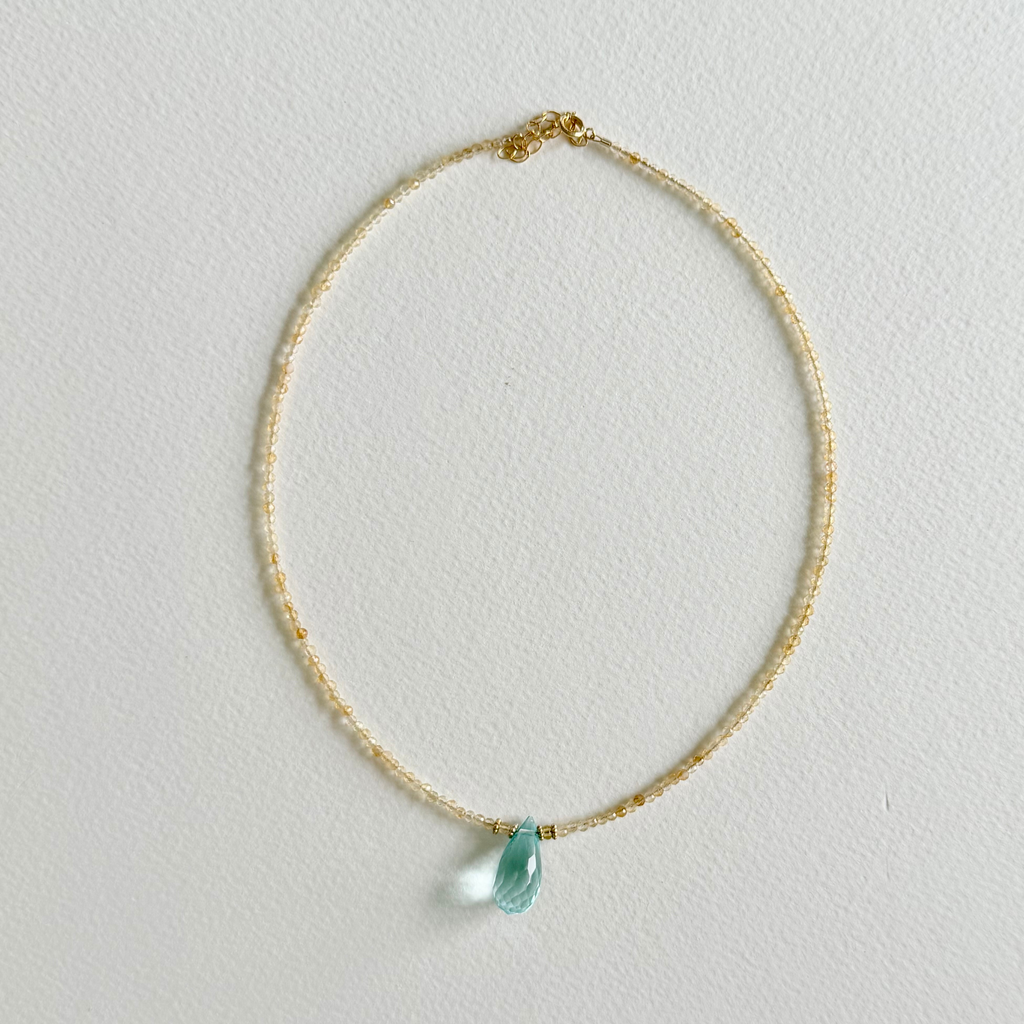 A delicate gold citrine beaded necklace with a small, faceted light blue aquamarinne teardrop gemstone pendant, displayed on a textured off-white background.