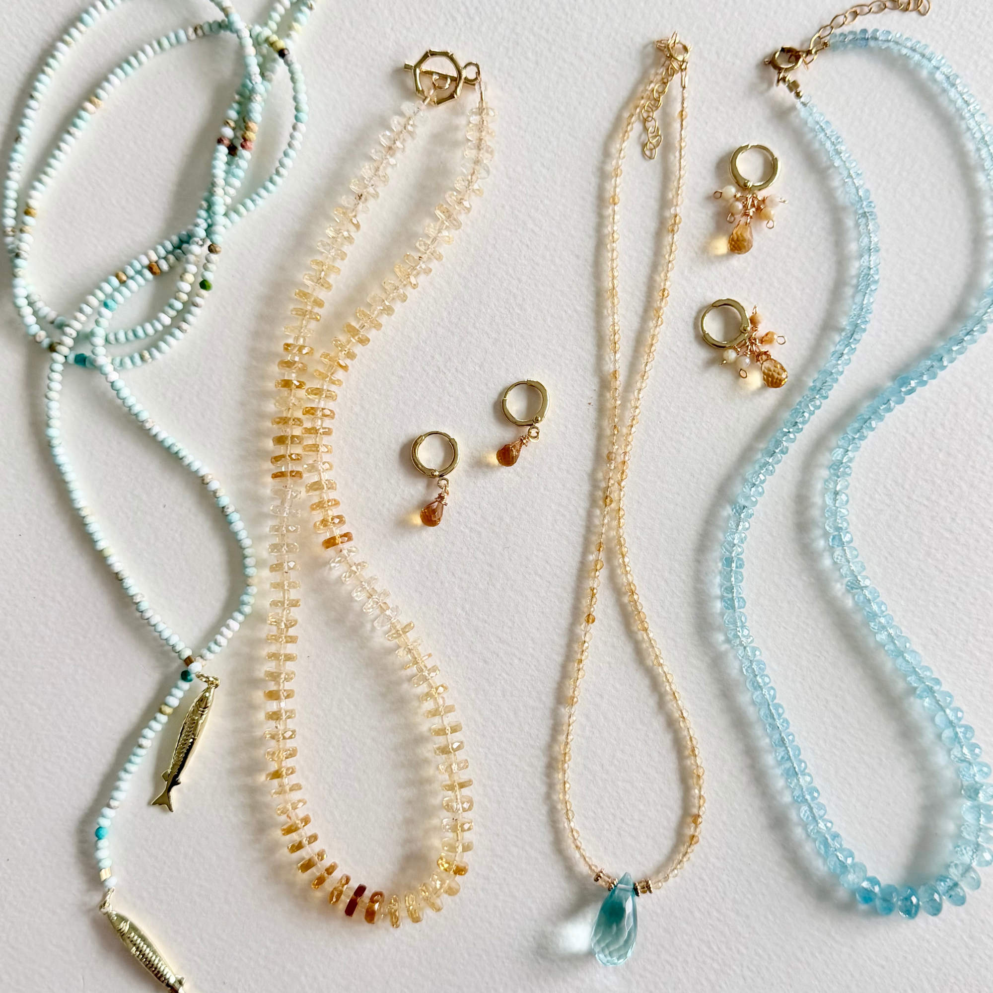 Four delicate beaded necklaces and matching drop earrings arranged on a light neutral background. The jewelry features pale aqua, cream, and light blue beads with small gold-toned metal findings, including a fish charm, bead accents, and dangling teardrop charms.