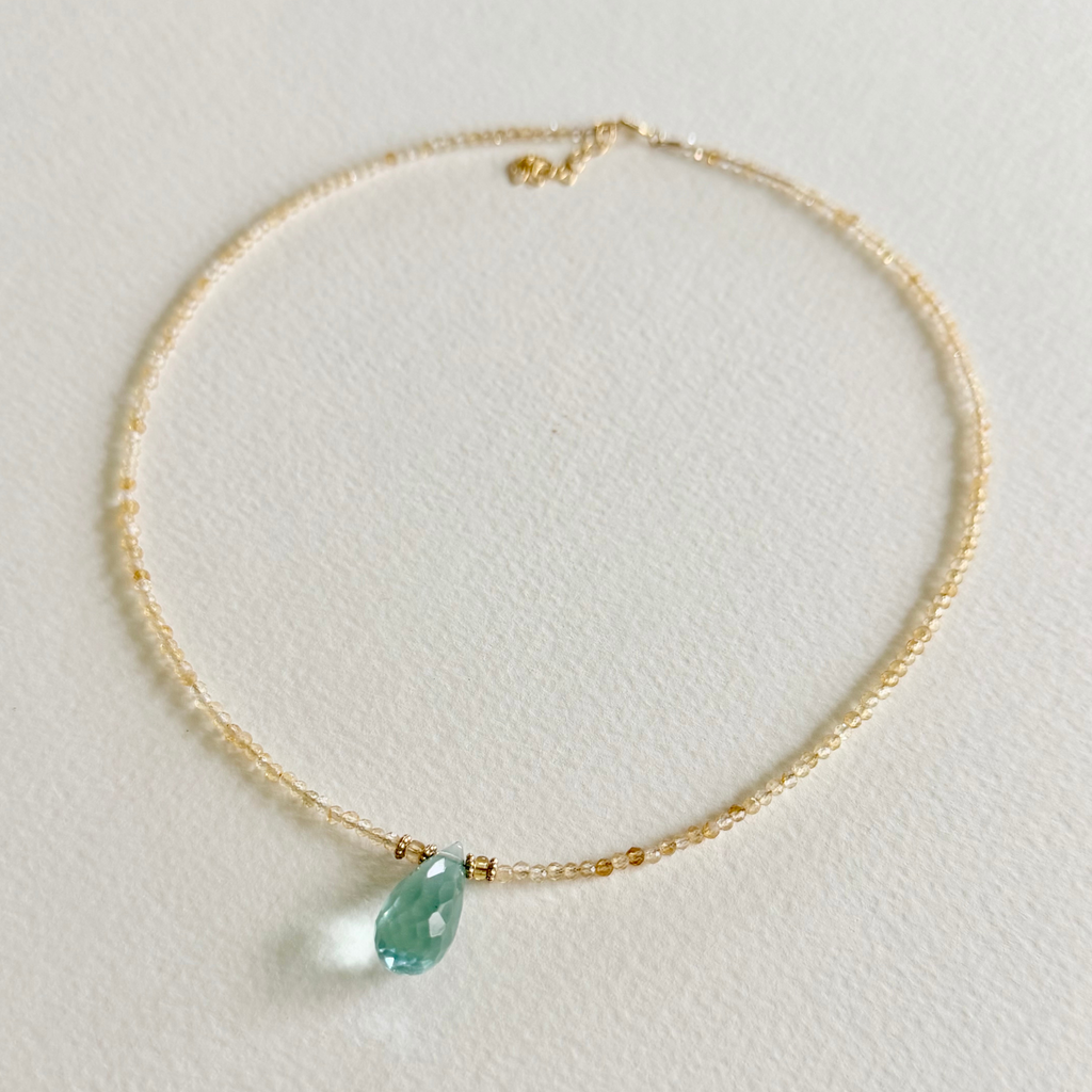 A delicate gold citrine beaded necklace with a small gold bead accent and a teardrop-shaped light blue aquamarine pendant, displayed on a textured off-white surface.