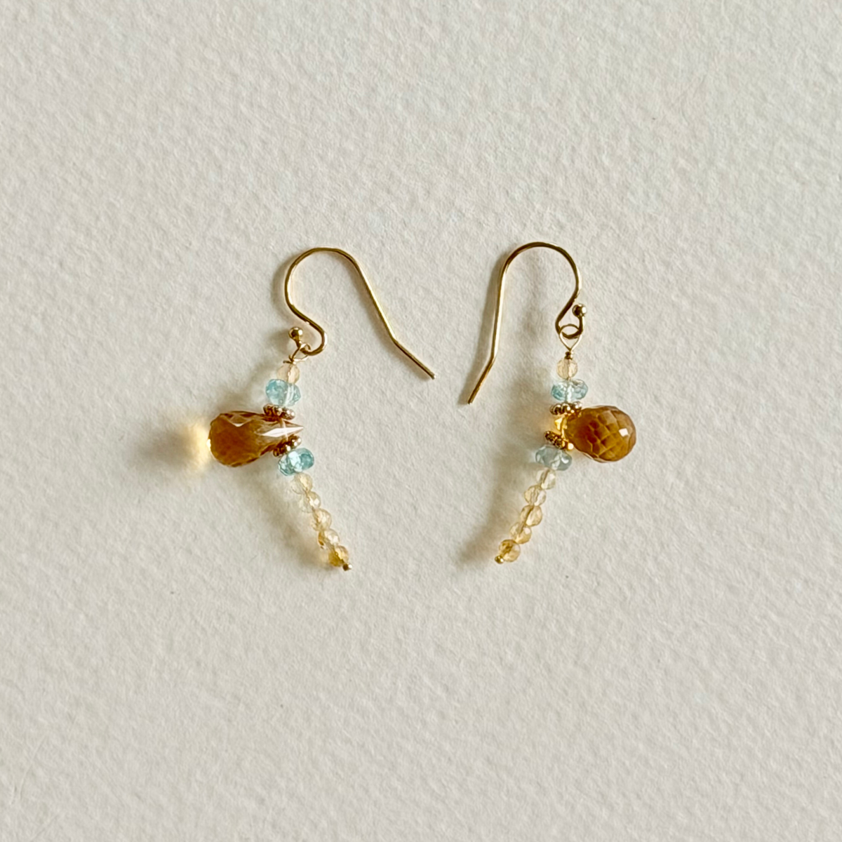 A pair of gold-tone dangle earrings with light blue natural zircon faceted beads, a larger amber-brown citrine teardrop bead, and small dangling gold beads, displayed on a textured off-white background.