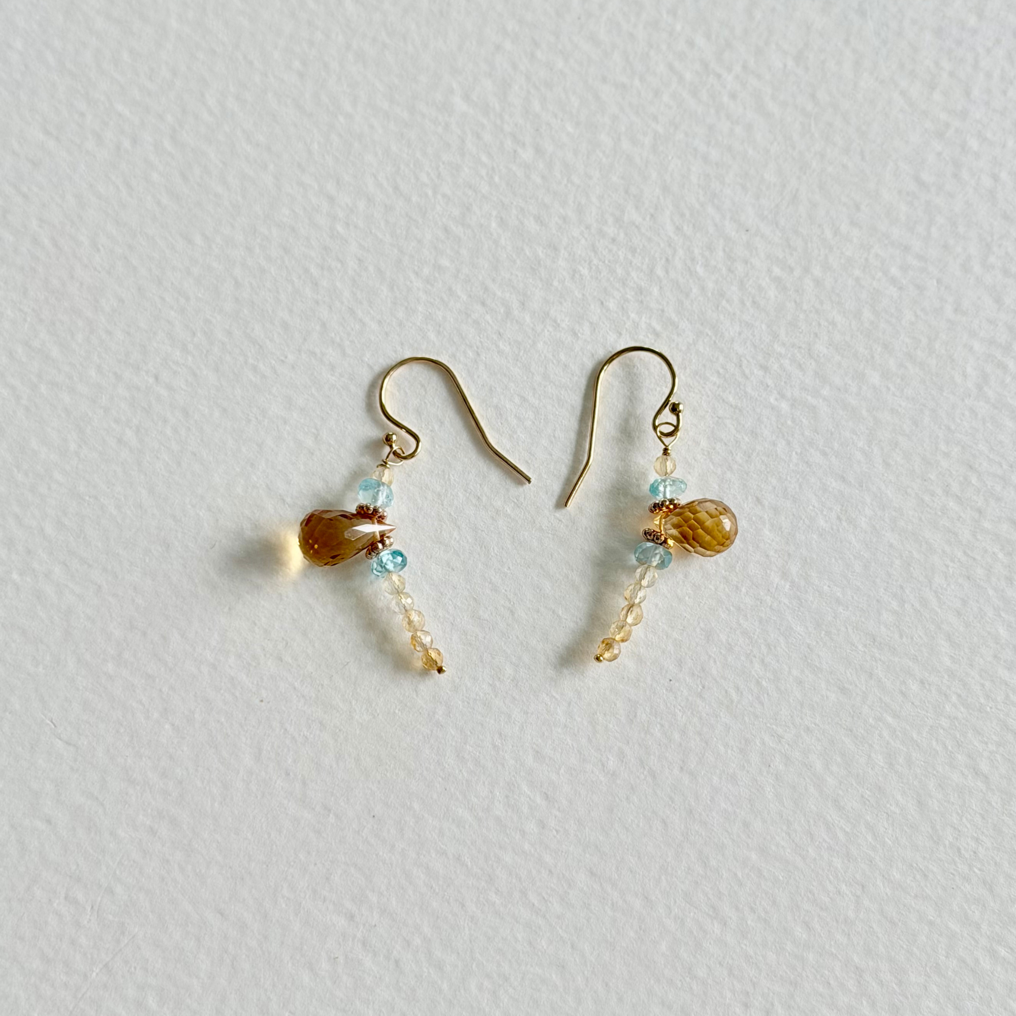 A pair of small gold-tone dangle earrings with citrine and light blue natural zircon beads, displayed on a textured off-white background.