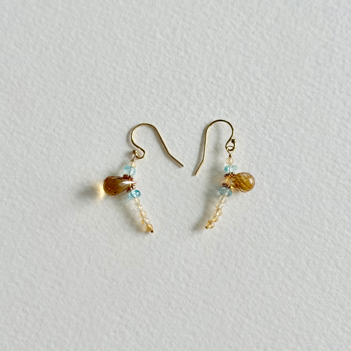 A pair of small gold-tone dangle earrings with citrine and light blue natural zircon beads, displayed on a textured off-white background.