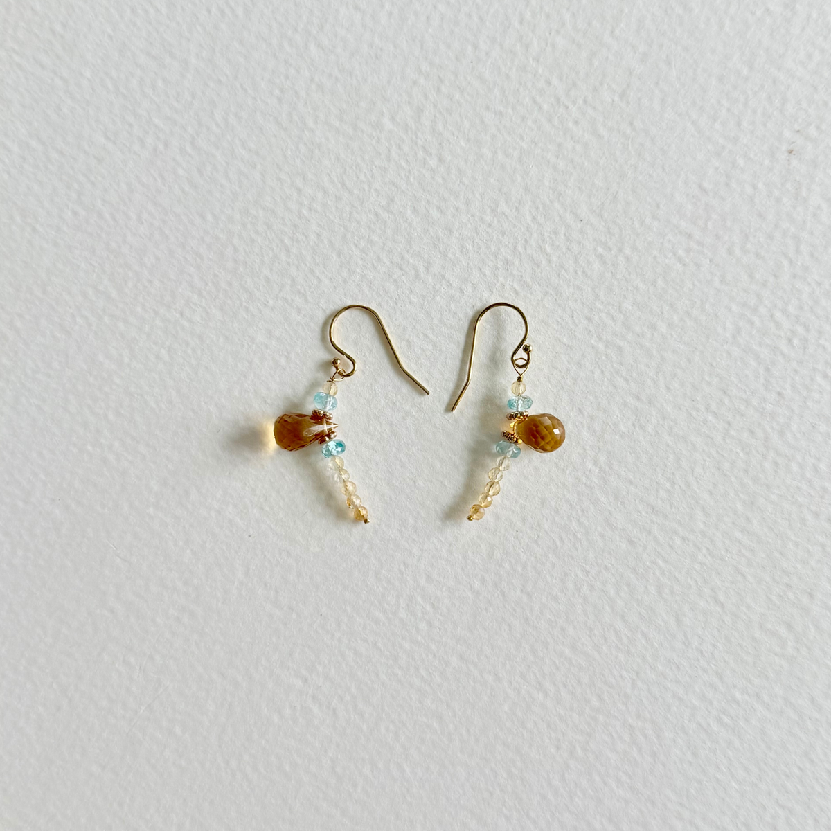 A pair of small gold-tone dangle earrings with citrine amber-brown oval beads, light blue natural zircon beads, and tiny gold bead accents, displayed on a textured off-white background.