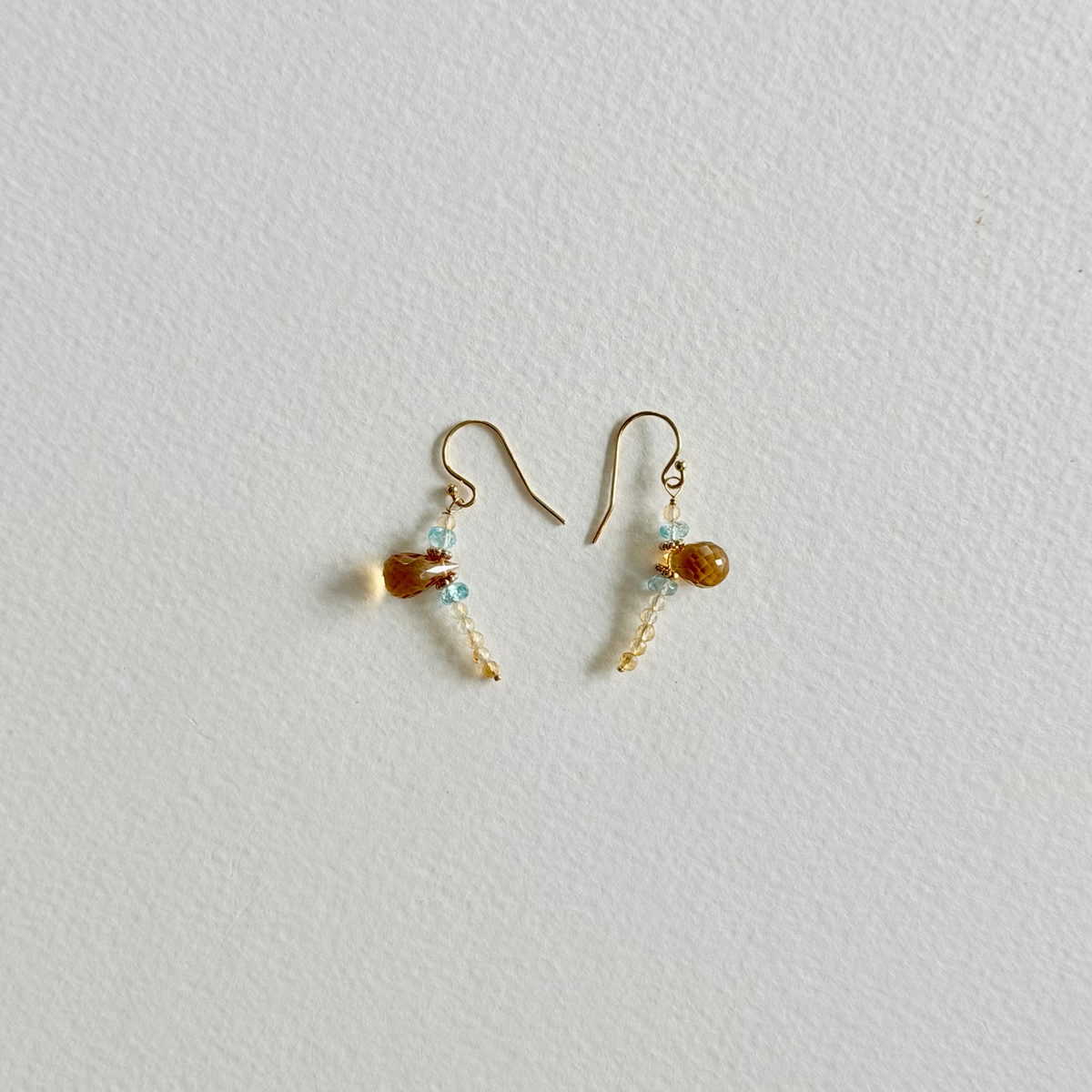 A pair of small gold-tone dangle earrings with amber-colored citrine beads and tiny light blue natural zircon beads, displayed on a textured off-white background.