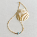 A delicate gold beaded citrine necklace featuring a central light blue natural zircon bead and small metallic accents, styled with a large cream-colored seashell on a textured off-white background.