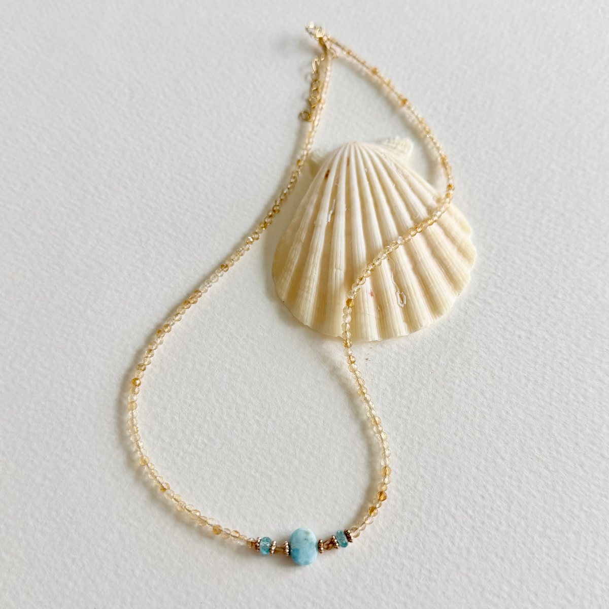 A delicate gold beaded citrine necklace featuring a central light blue natural zircon bead and small metallic accents, styled with a large cream-colored seashell on a textured off-white background.