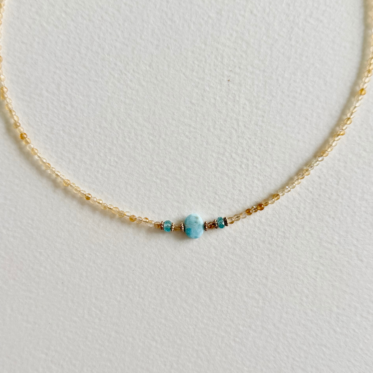 A delicate gold beaded necklace featuring a central larimar bead flanked by small gold and natural zircon spacer beads, displayed on a textured off-white background.