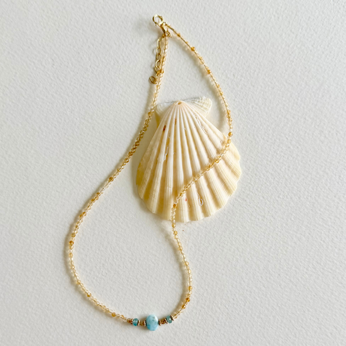 A citrine gold-toned beaded necklace styled with a large cream-colored seashell pendant and a small turquoise bead accent, displayed on a textured off-white background.