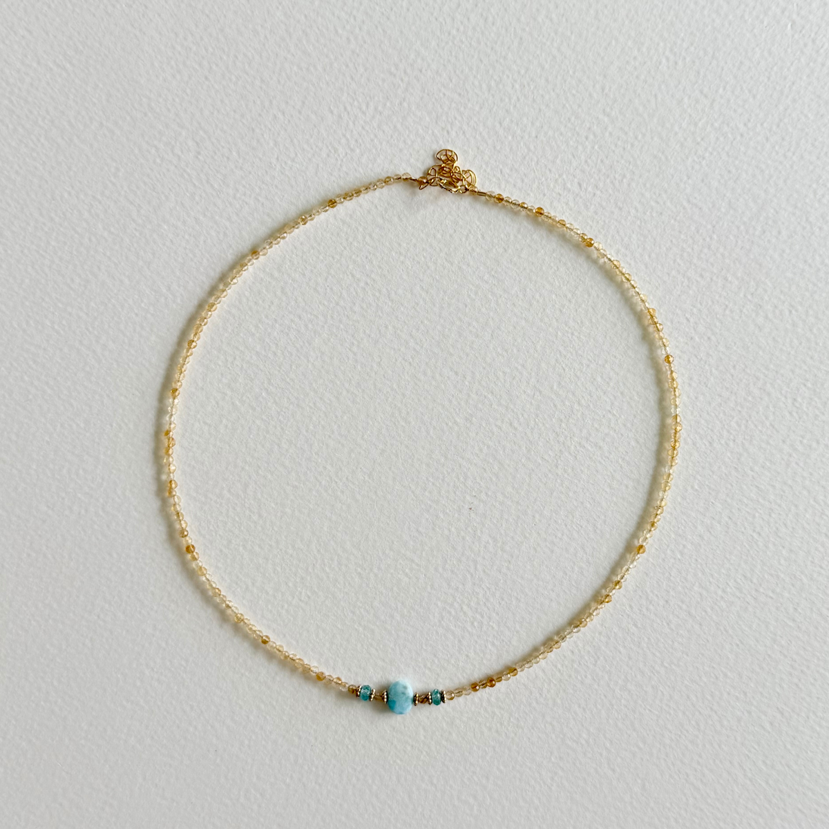 A delicate gold citrine beaded necklace with a small larimar bead and tiny gold daisy spacer beads, displayed on a textured white background.