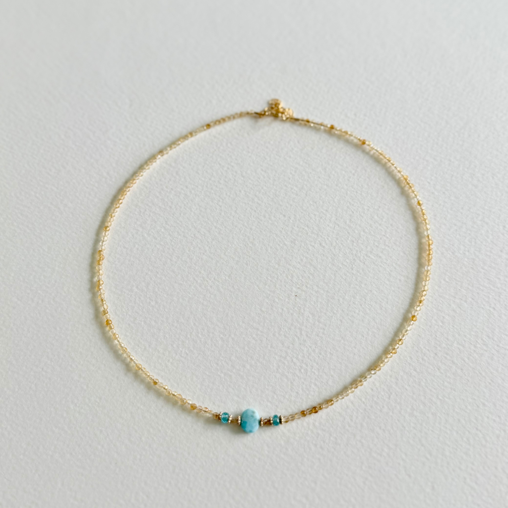 A delicate gold beaded citrine necklace with a small larimar bead and tiny gold accent beads, displayed on a textured white surface.