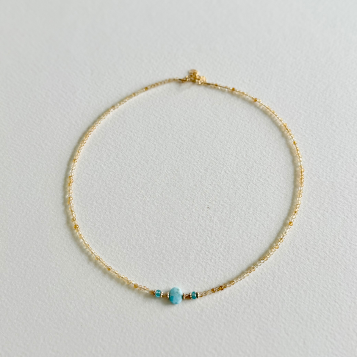 A delicate gold beaded citrine necklace with a small larimar bead and tiny gold accent beads, displayed on a textured white surface.