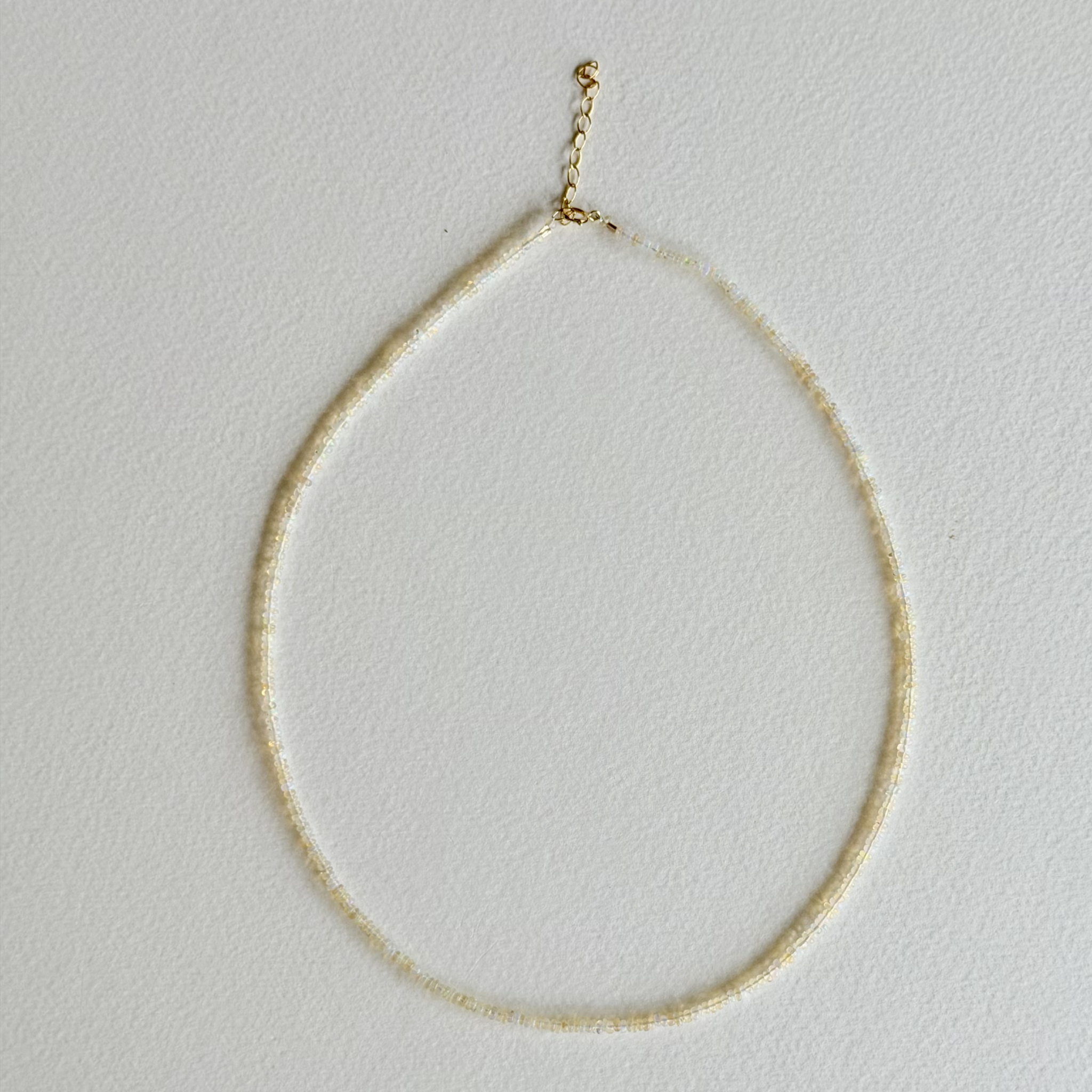A delicate beaded necklace with small, round cream-colored beads and a gold-toned clasp, displayed against a textured off-white background.