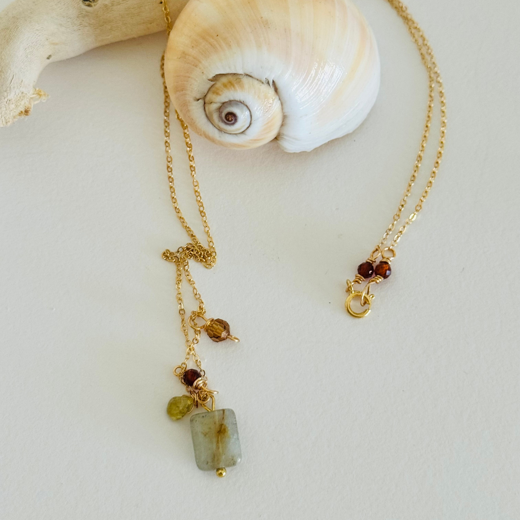 Simple chain necklace with one each of aventurine, peridot, and garnet hessonite beads in warm fall coastal colors displayed draped over a creamy spiral seashell and bleached driftwood. Two garnet beads frame the clasp.