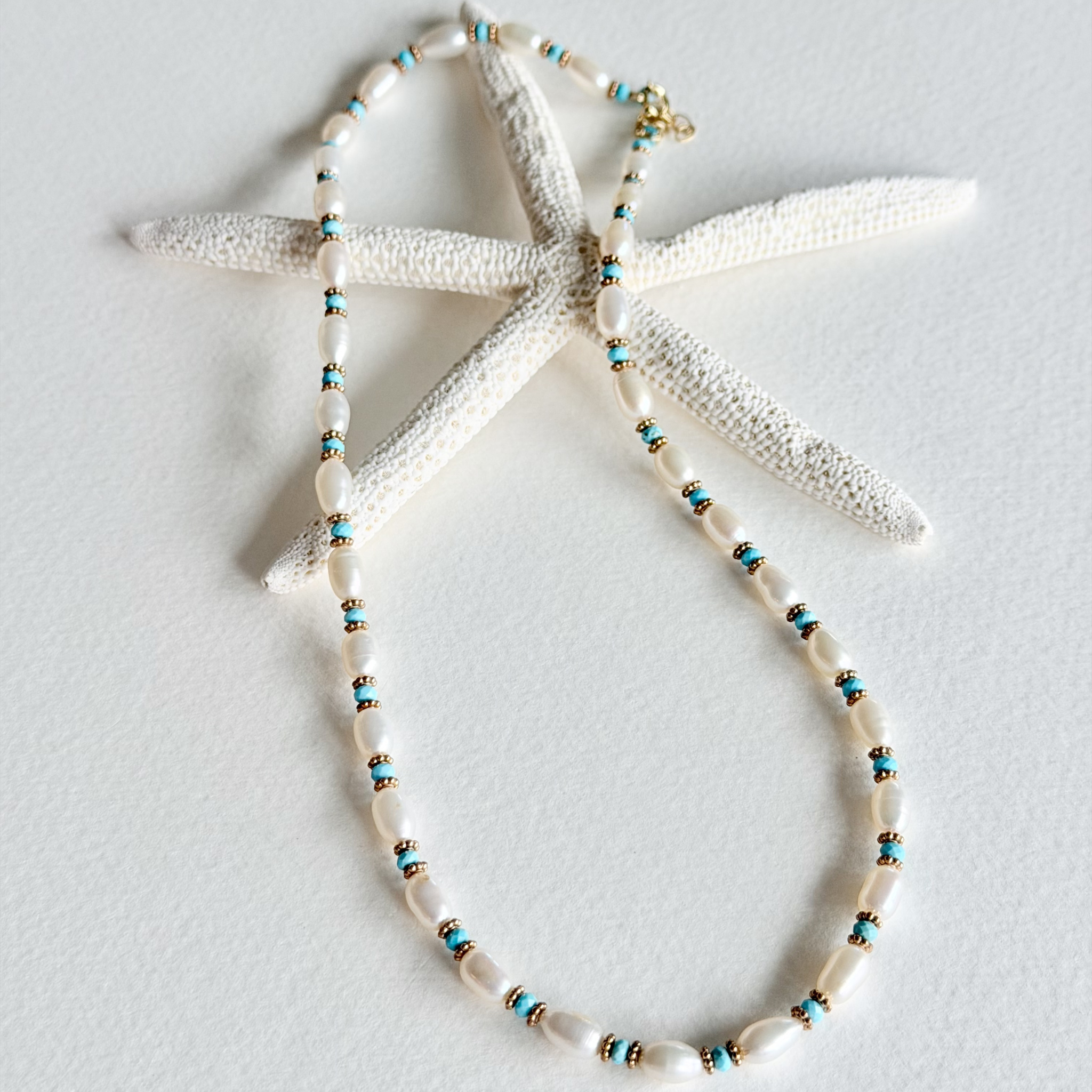 A beaded necklace with white shell-like beads and small turquoise and gold spacer beads, displayed on a white starfish prop against a light gray background.