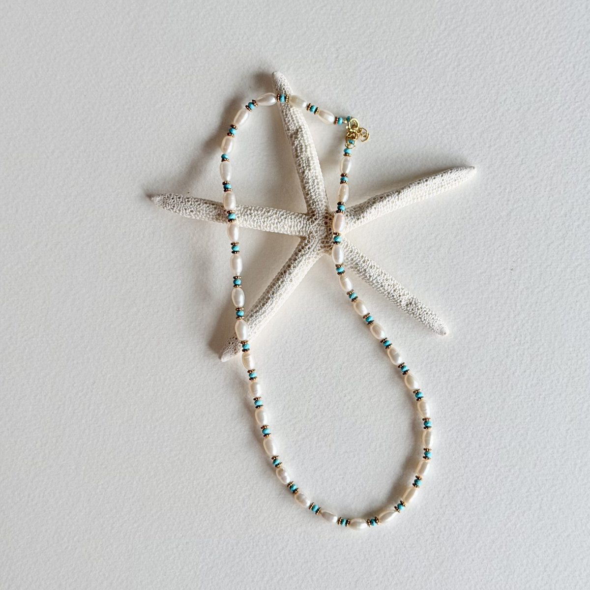 A beaded necklace with white shell-like beads and small turquoise and gold accents, displayed on a white starfish prop against a light gray background.