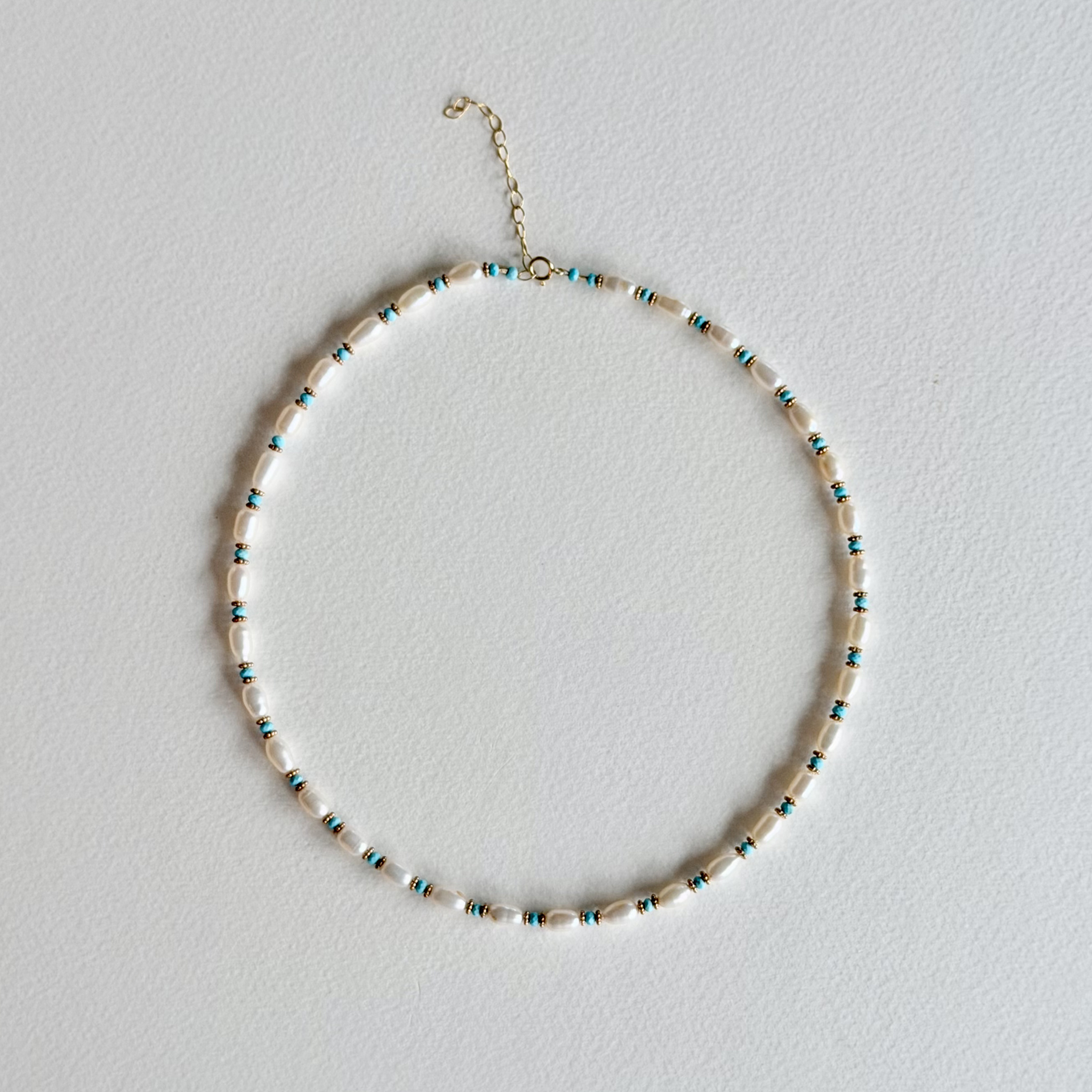 A delicate beaded necklace with white oval beads and small turquoise and gold spacer beads, displayed on a textured white background.