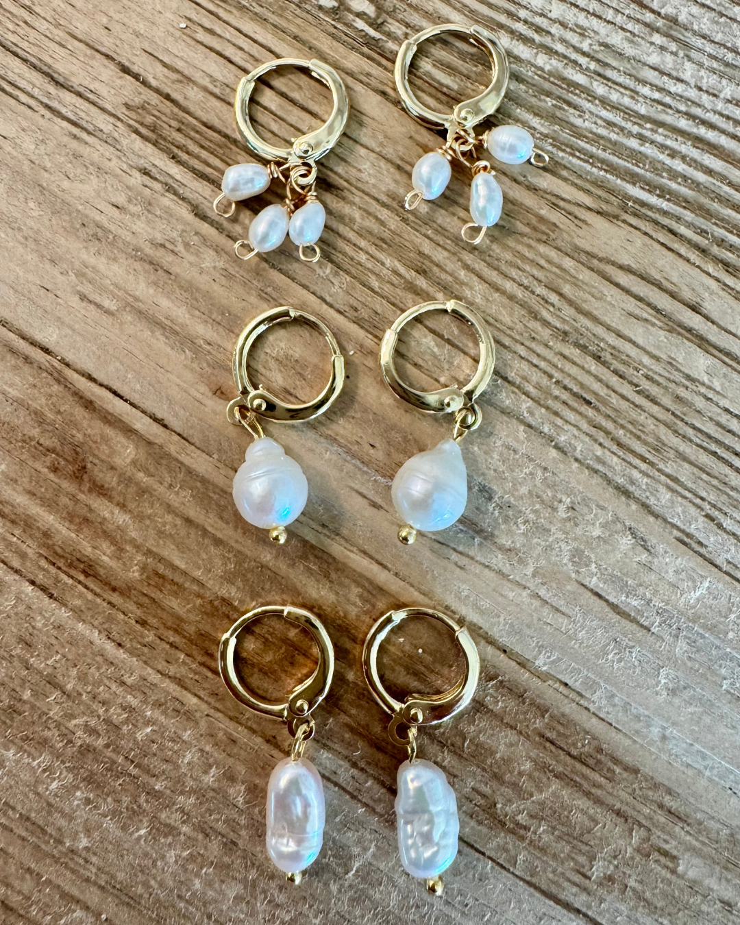 Set of three huggie earrings collection. From top to bottom, a pair of earrings with three oval pearls, a pair of earrings with a pear-shaped pearl, and a pair of earrings with a single oblong freshwater pearl on each hoop all handcrafted with 14k gold-filled wire huggie clasp. 