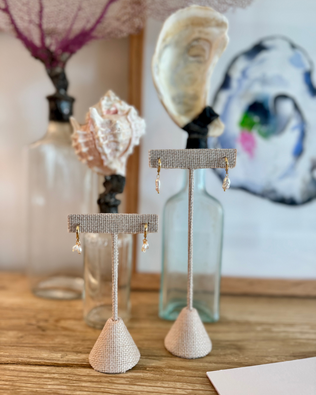 Huggie earrings displayed on linen burlap jewelry holders in from of seashell display bottles and oyster art print.