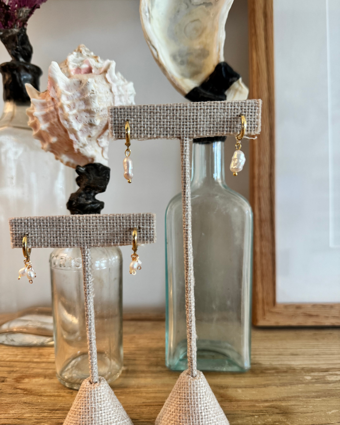 Two pairs of huggie earrings displayed on linen burlap jewelry stands in front of seashell and oyster display glass jar bottles.
