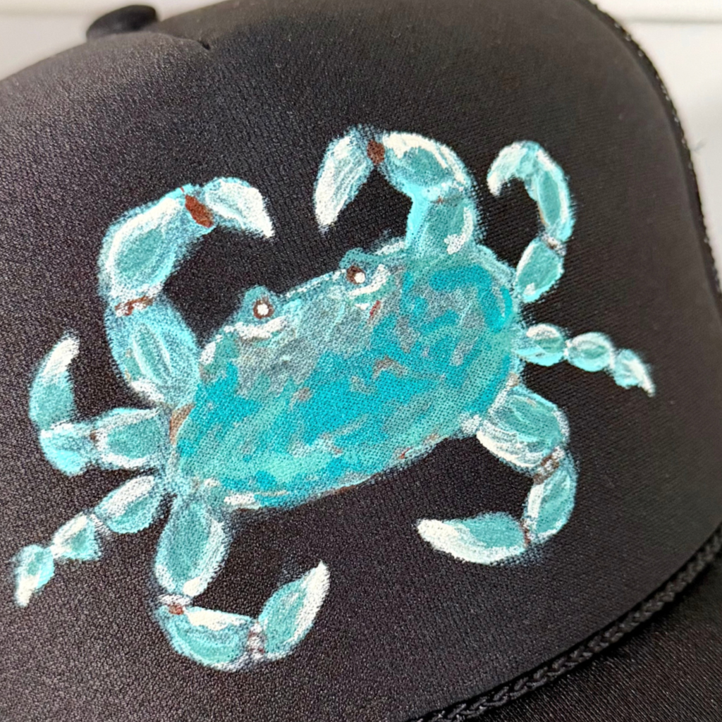 A close-up of a black trucker-style cap featuring a hand-painted turquoise blue crab on the front panel.