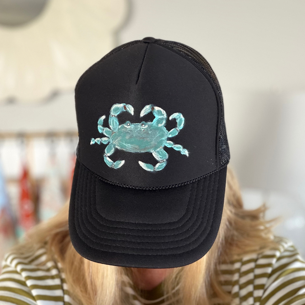 A person wearing a black trucker hat with a light blue embroidered crab on the front, photographed indoors with a blurred background.