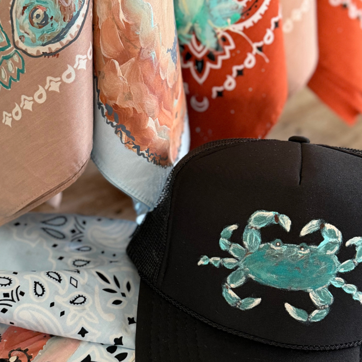 A black trucker hat with a turquoise crab graphic on the front, styled in front of a display of colorful patterned scarves.