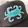 A close-up of a black trucker-style cap featuring a hand-painted turquoise blue crab on the front panel.