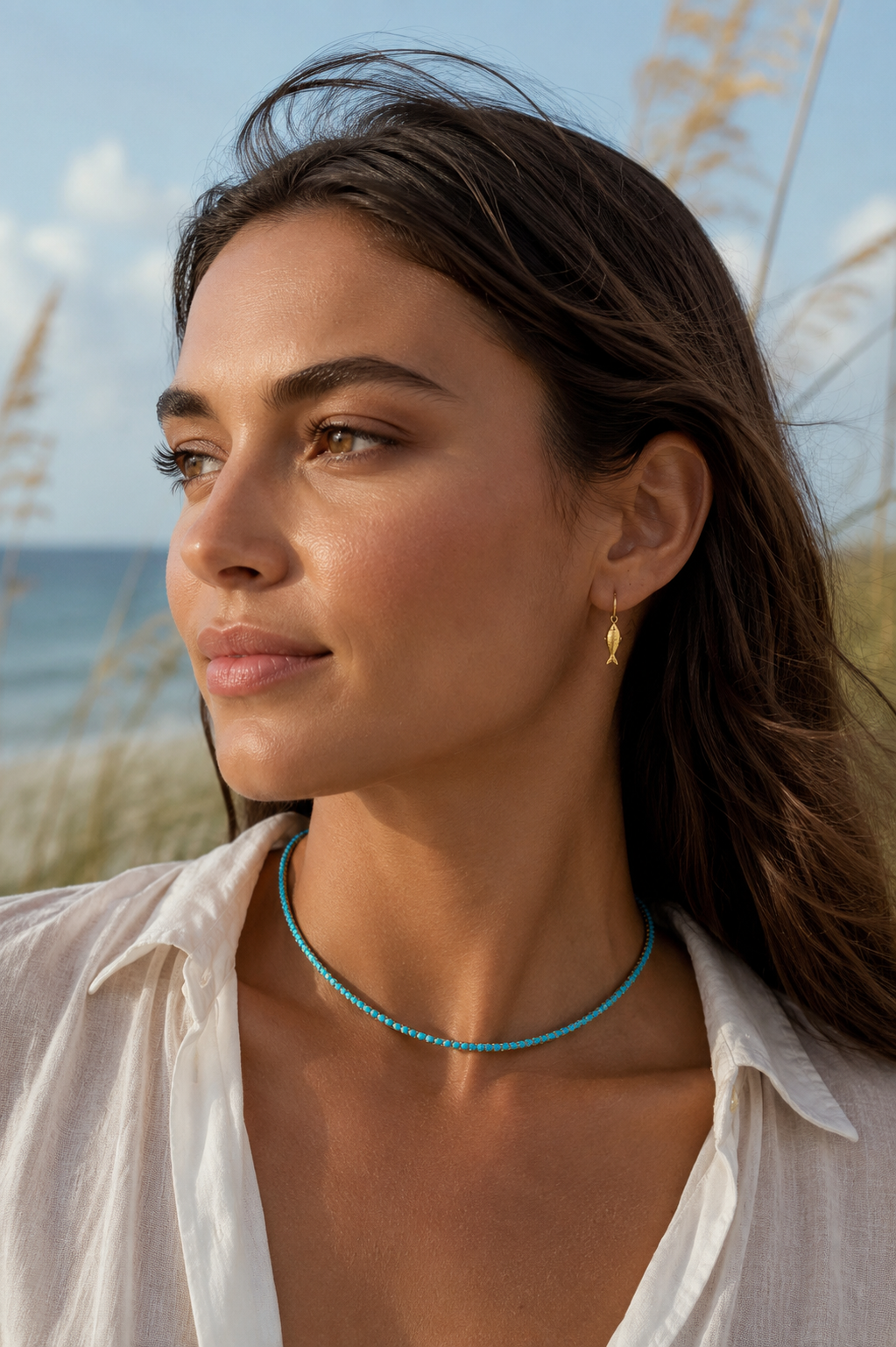 A woman outdoors near the beach wearing a thin turquoise beaded necklace and a small gold hoop earring, with a white shirt and blurred reeds and ocean in the background.
