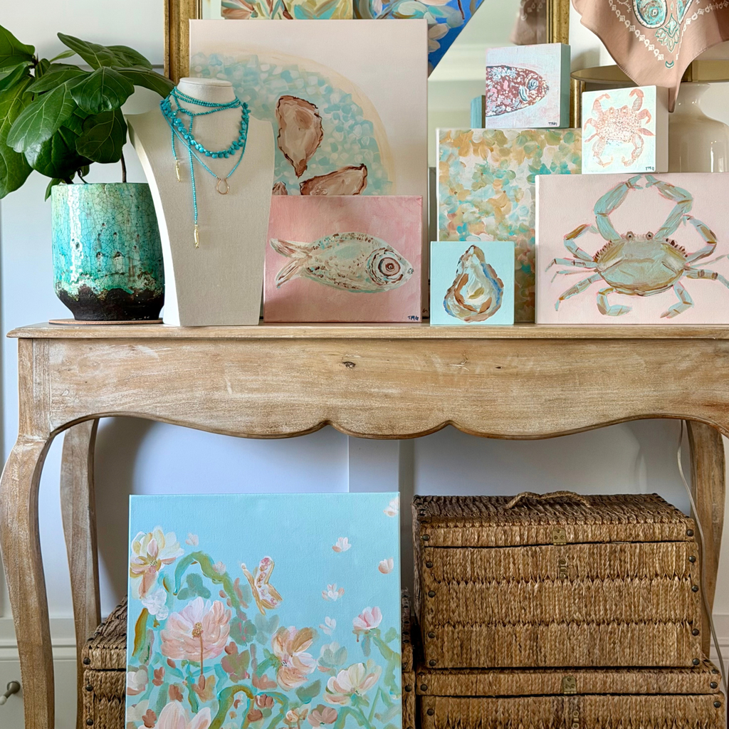A styled interior scene showing a wooden console table with several pastel art blocks and paintings featuring sea life and floral designs. A turquoise ceramic planter with green leaves sits on the left, and woven storage baskets are stacked underneath.