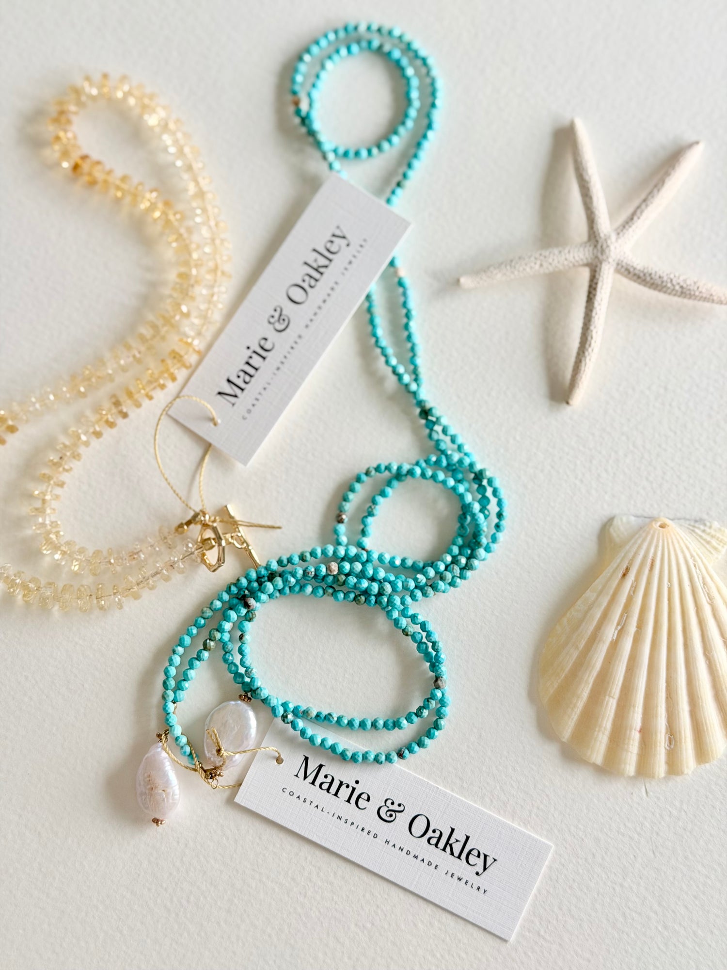 Two beaded strands from Marie & Oakley jewelry displayed on a white background with decorative seashells. One strand is a translucent pale yellow bead necklace, and the other is a turquoise blue beaded bracelet/necklace with a white teardrop shell charm.