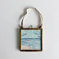 A small square framed painting of a beach landscape with blue water and sky, hanging from a brown leather strap against a white wall.