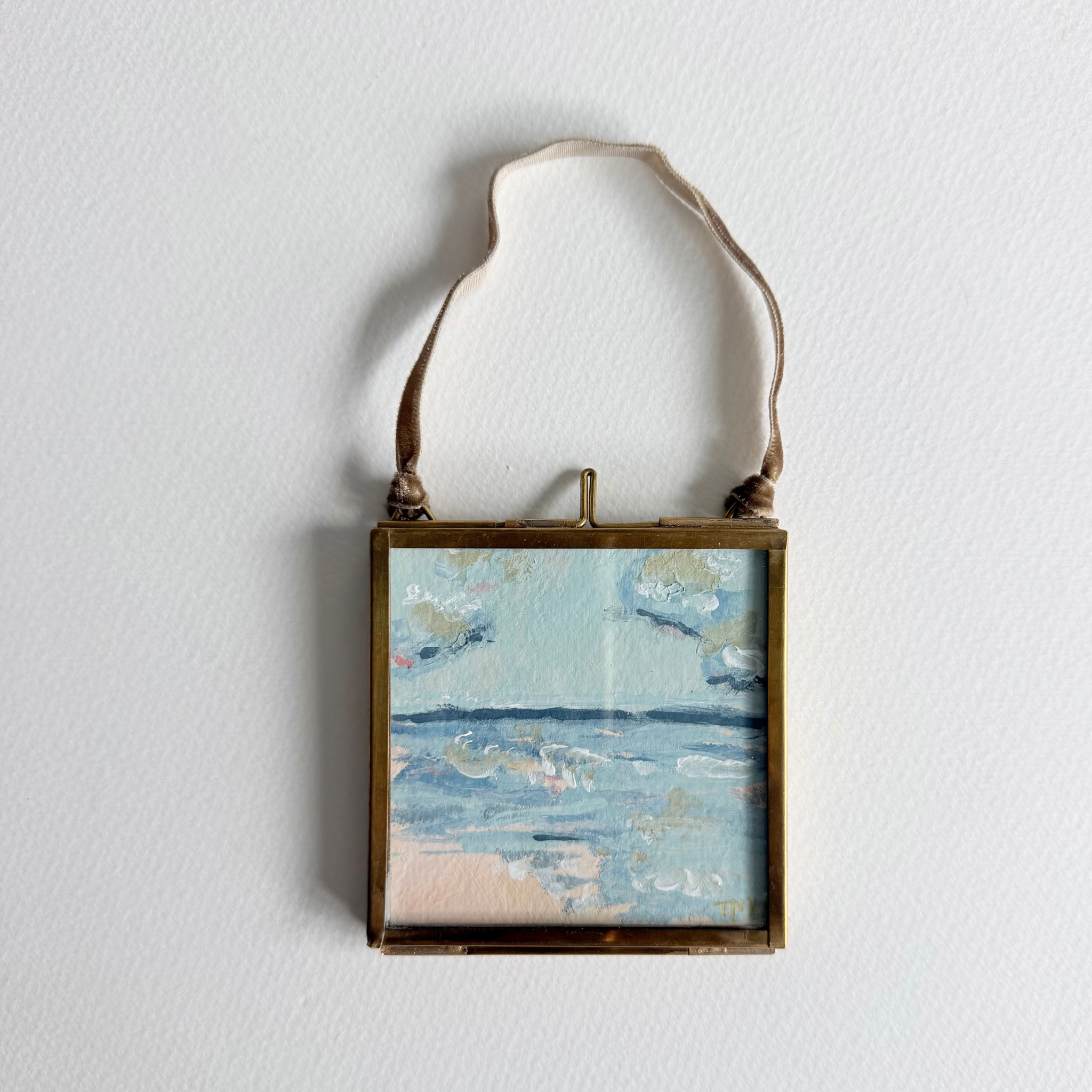 A small square framed painting of a beach landscape with blue water and sky, hanging from a brown leather strap against a white wall.