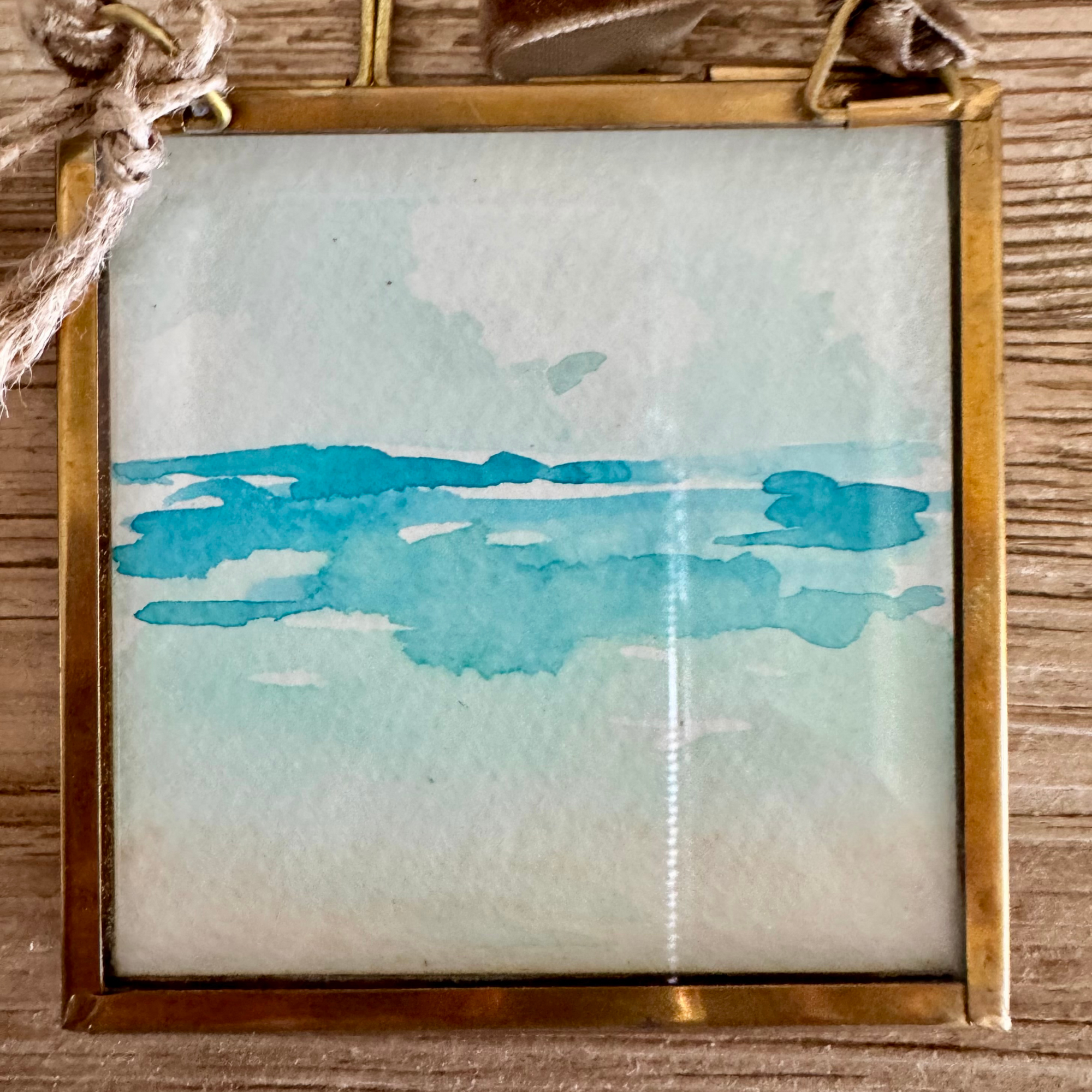 Watercolor of seascape in blue and tan. Framed in brass square glass frame and tied with brown velvet ribbon to hang as an ornament.