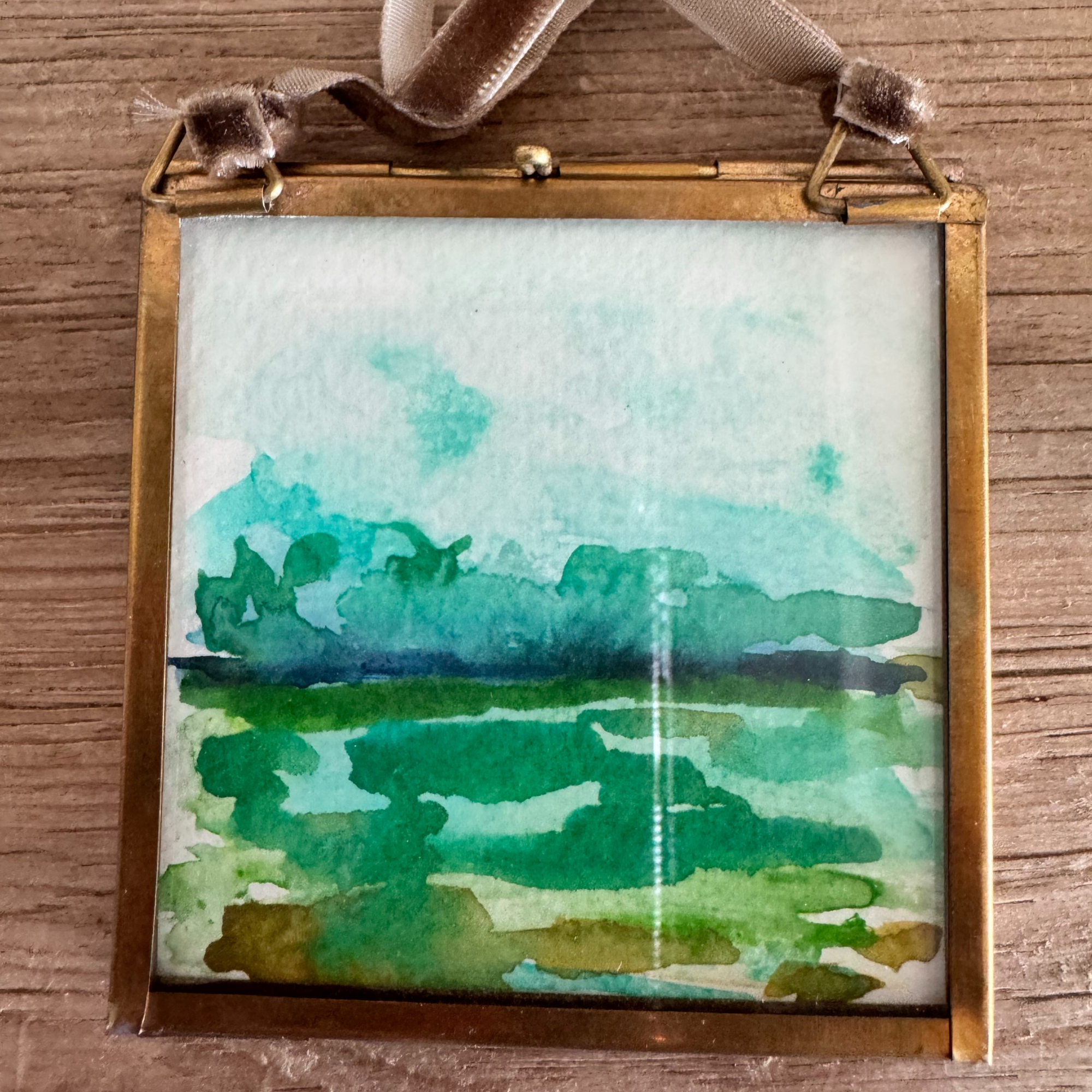 Watercolor of seascape in silver, green, and navy blue. Framed in brass square glass frame and tied with brown velvet ribbon to hang as an ornament.