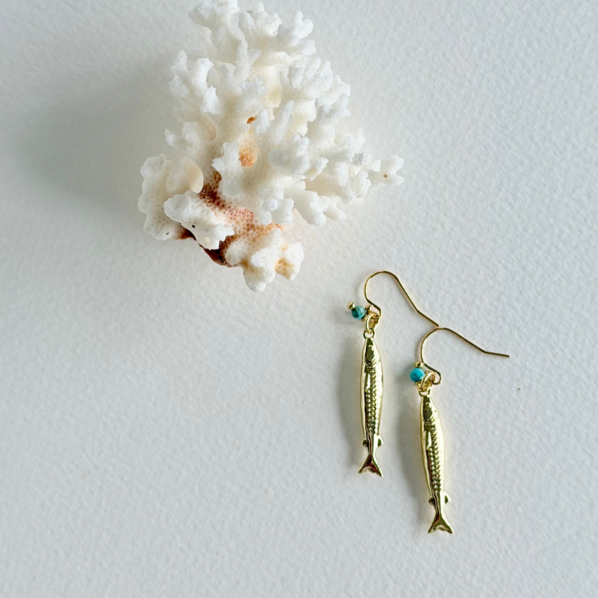 Gold fish-shaped earrings with turquoise beads on a white background with coral.