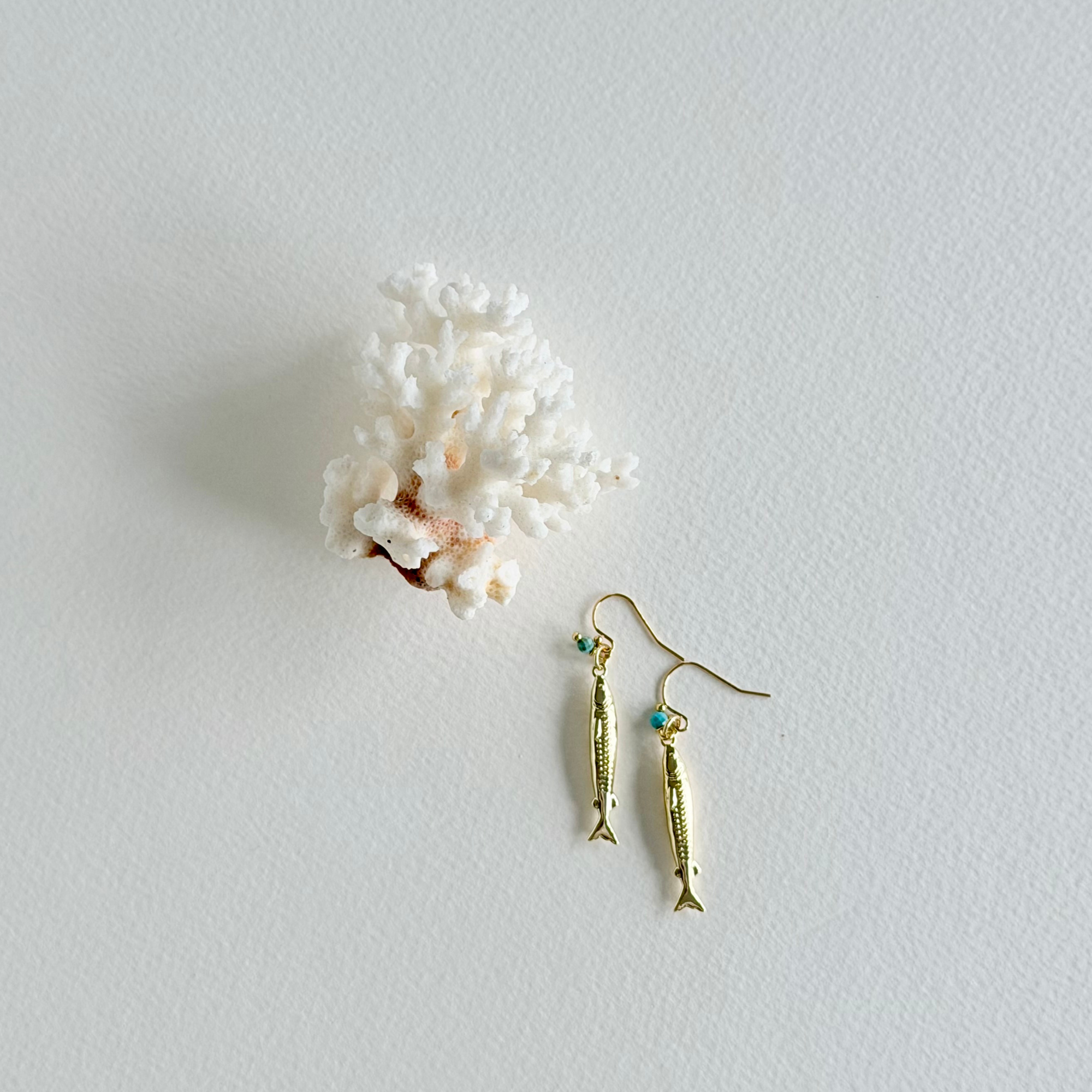Gold fish-shaped earrings with turquoise stones on a light gray background with white coral.