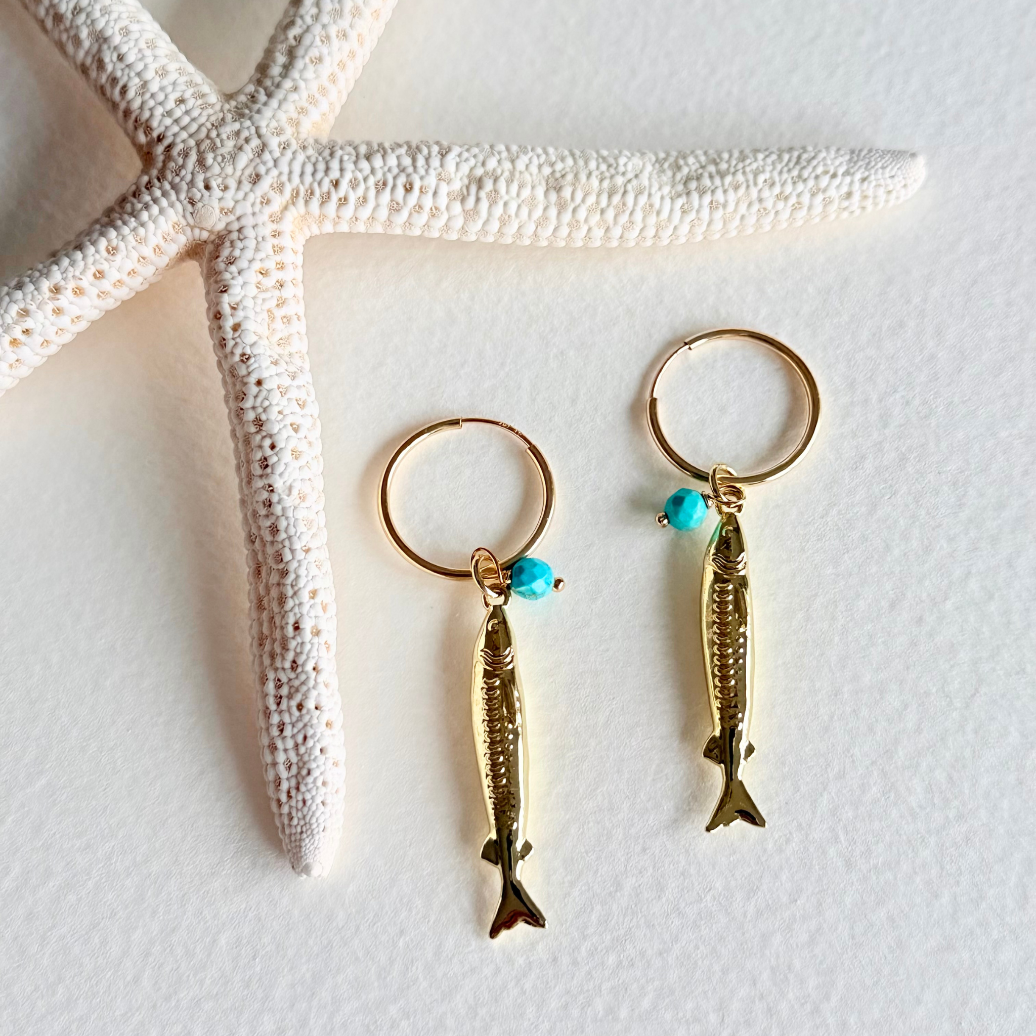 Gold fish-shaped earrings with turquoise beads on a white surface with a starfish.