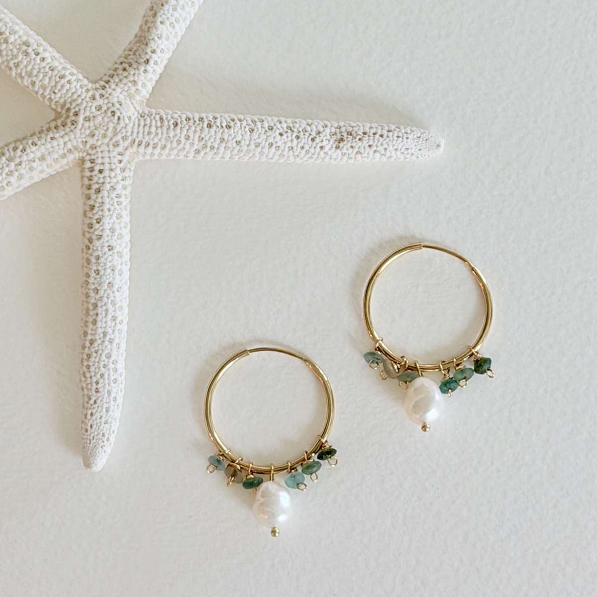 Gold hoop earrings with green beads and pearls on a white surface with a starfish.